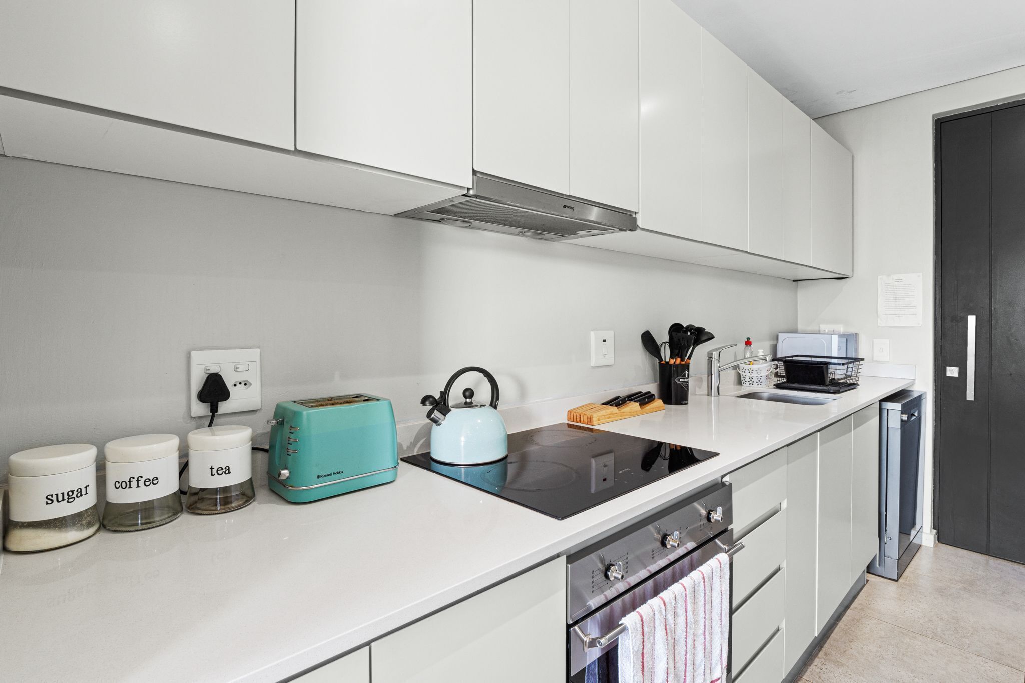 A modern kitchen with white cabinets, a black stove, a teal kettle, and labeled containers for sugar, coffee, and tea.