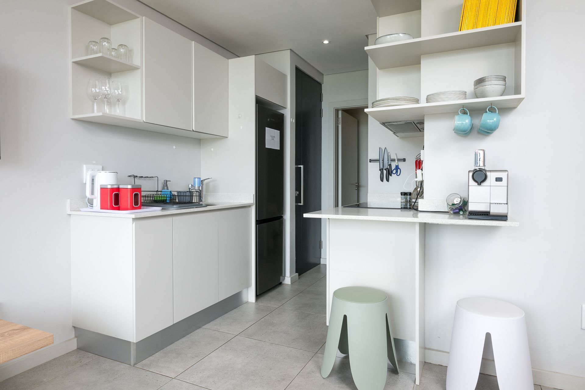 A modern kitchen with white cabinetry, a built-in oven, and a coffee machine. There are two stools and a small table.