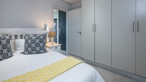 Un dormitorio con cama, dos mesitas de noche, lámpara, armario y puerta que da a otra habitación.