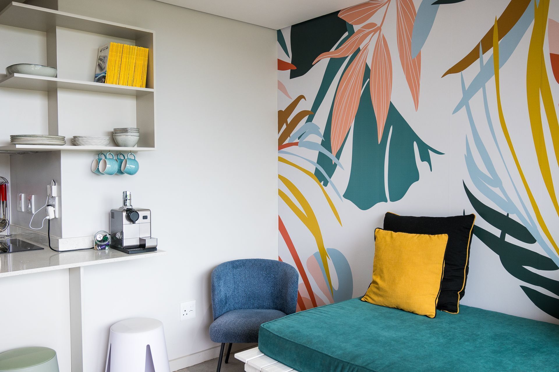 A modern bedroom with a bed, a chair, a desk with shelves, and a coffee machine.