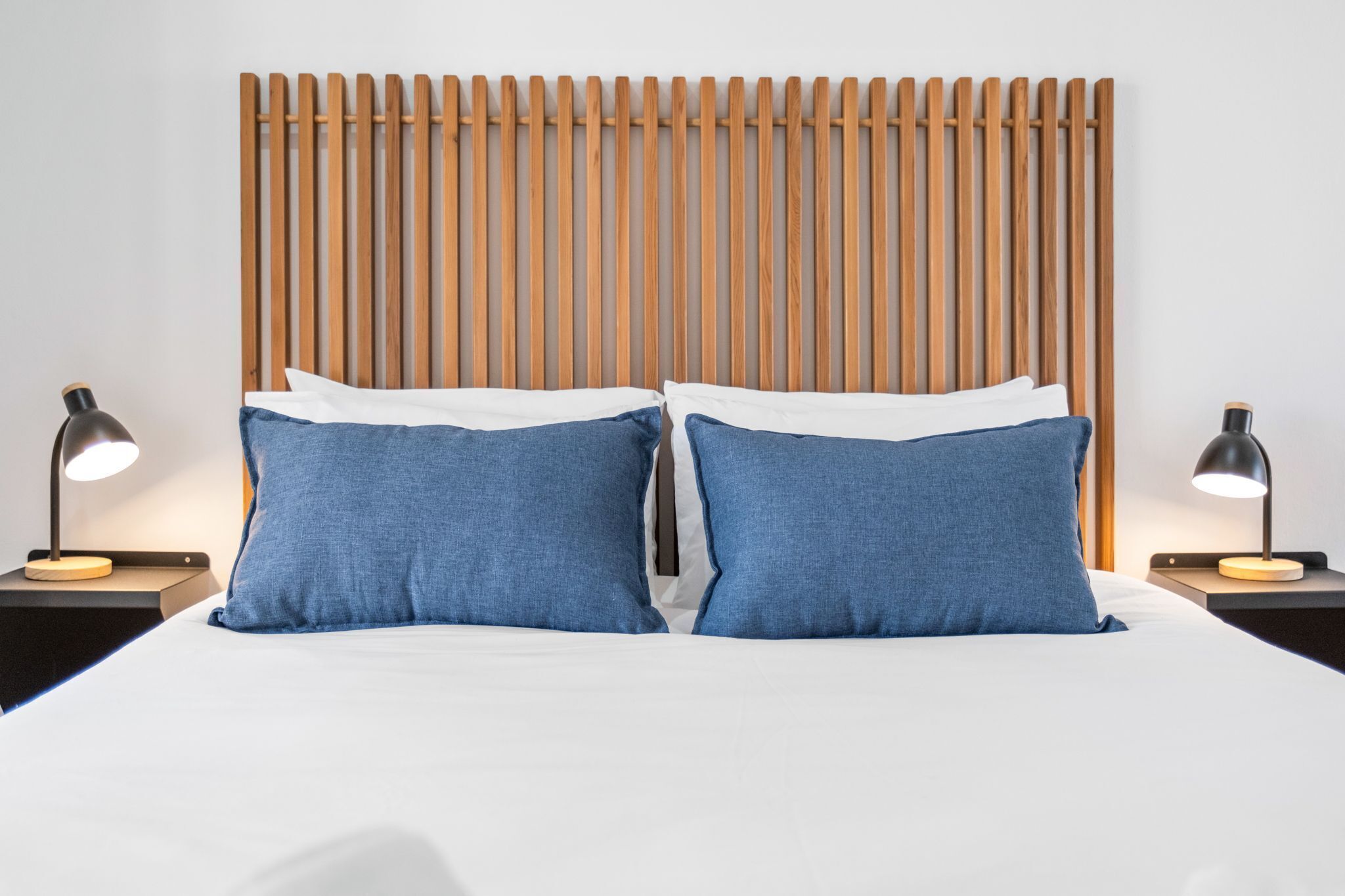 A neatly made bed with white and blue bedding, flanked by two bedside tables with lamps, against a backdrop of a wooden slat headboard.