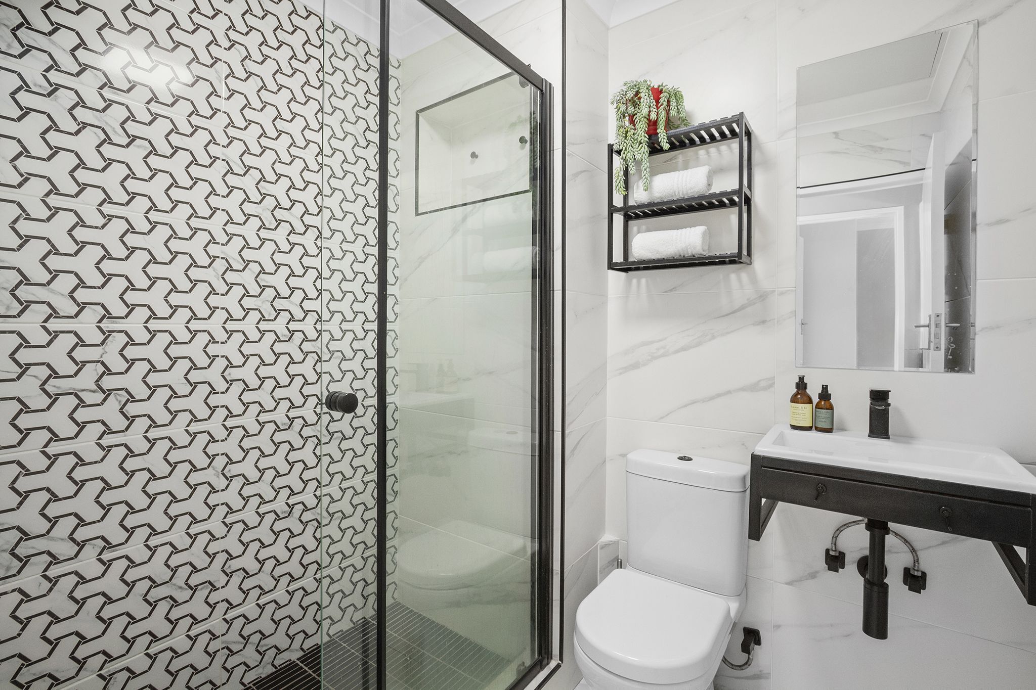 A modern bathroom with a geometric patterned wall, a glass shower enclosure, a white toilet, a black vanity with a mirror, and a shelf with toiletries.