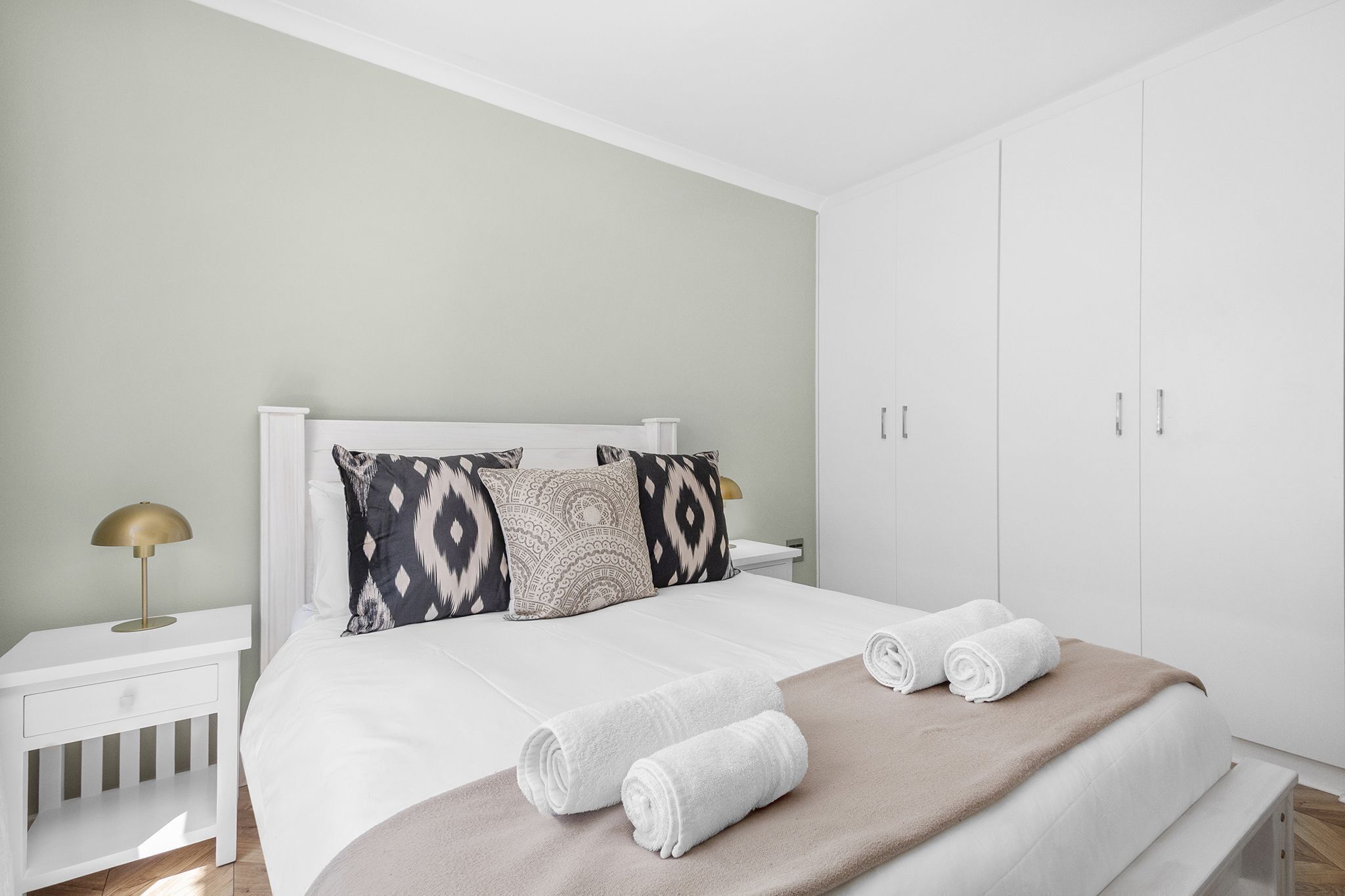 A neatly made bed with white linens, two rolled towels, and patterned pillows.