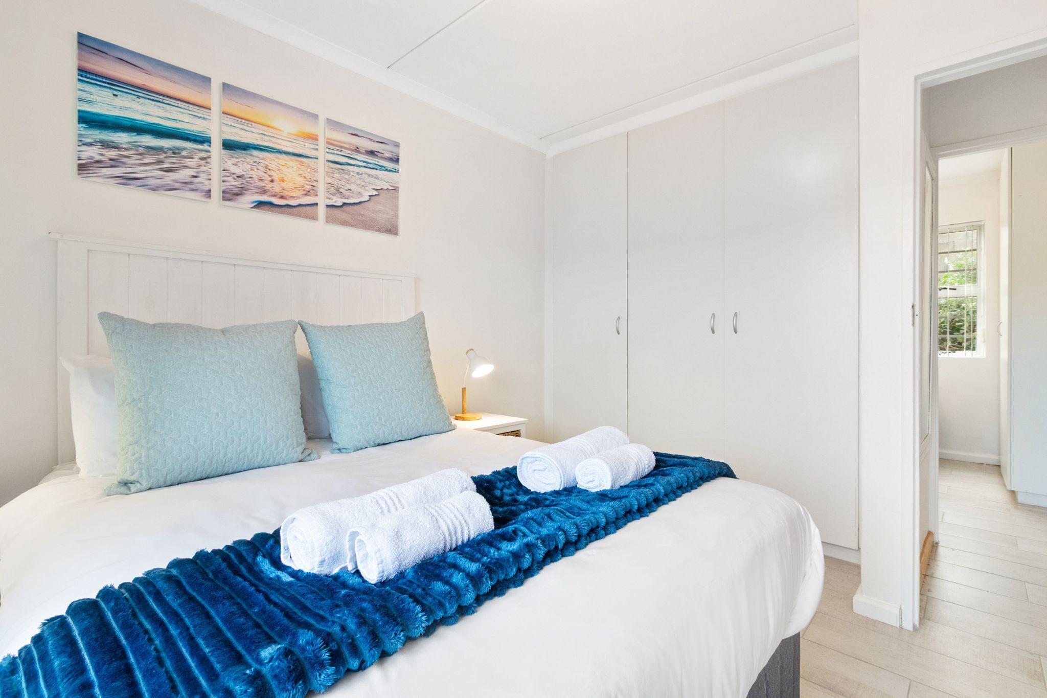 A hotel room with a bed, a nightstand, a blue blanket, and a view of the sea through the window.