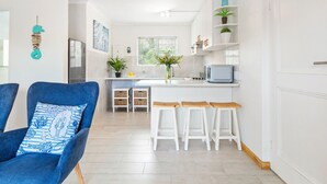 A modern kitchen with white cabinetry, a central island with stools, a refrigerator, and a potted plant. - Big Bay Beach Club 153 (Cape Town)