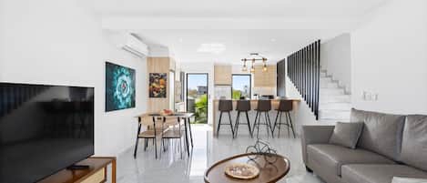 A modern living room with a flat-screen TV, a sofa, a coffee table, and a dining area with chairs and a table.
