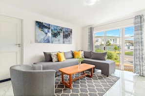 A modern living room with a grey sofa, a wooden coffee table, and a patterned rug. There are abstract paintings on the wall and a view of the outside through large windows. - Big Bay Beach Club 137 (Cape Town)