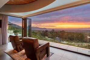 Ein modernes Zimmer mit einer großen Glasdoor, die auf einen Balkon mit herrlichem Bergblick und Sonnenuntergang führt.