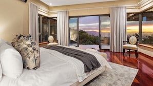 Une chambre spacieuse avec un grand lit, un fauteuil et une vue sur le coucher de soleil, grâce à de grandes fenêtres du sol au plafond.
