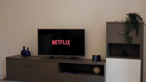 Ett modernt vardagsrum med en platt-TV som visar Netflix-logotypen, en grå TV-bänk med två blå vaser och en krukväxt på en vit hylla.