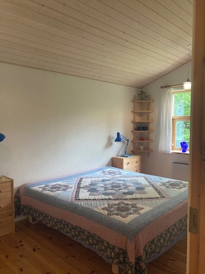 Ein Schlafzimmer mit Bett, Nachttisch, Kommode, Schreibtisch, Lampe, Fenster mit Blick und einem Regal mit Gegenständen.