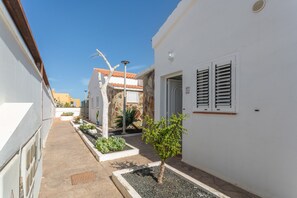 Une ruelle étroite avec un petit arbre, un bâtiment blanc avec des volets et, au fond, un mur de pierre surmonté d’un toit en tuiles.