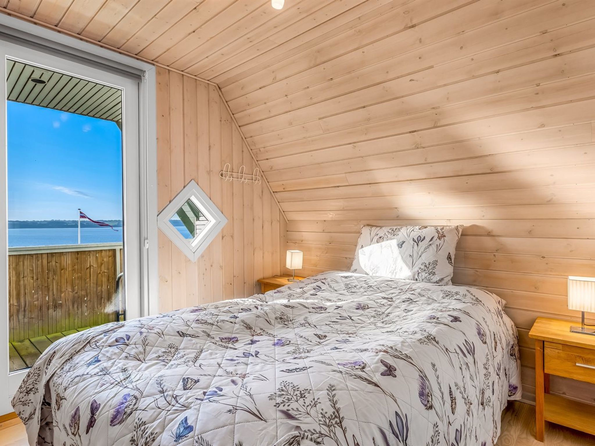 A bedroom with a wooden sloped ceiling, a bed with a floral quilt, a nightstand with a lamp, and a view of the sea through a window.