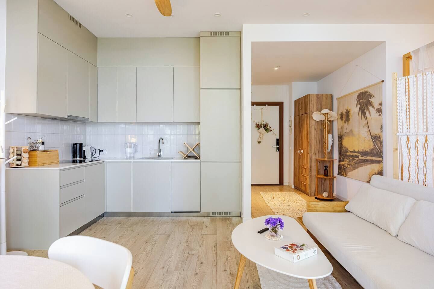 Una cocina moderna con armarios blancos, una cafetera y un lavamanos. Hay un sofá blanco y una mesa de centro con un libro y un jarrón de flores.