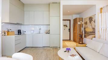 Una cocina moderna con armarios blancos, una cafetera y un lavamanos. Hay un sofá blanco y una mesa de centro con un libro y un jarrón de flores.