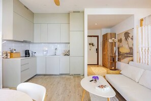 Una cocina moderna con armarios blancos, una cafetera y un lavamanos. Hay un sofá blanco y una mesa de centro con un libro y un jarrón de flores.