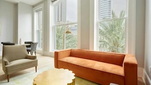A modern living room with a beige sofa, a round coffee table, and a view of palm trees through large windows. - Walk to Bourbon St, Riverfront Park, French Qtr | Canal St Stay | The Harlequin (New Orleans)