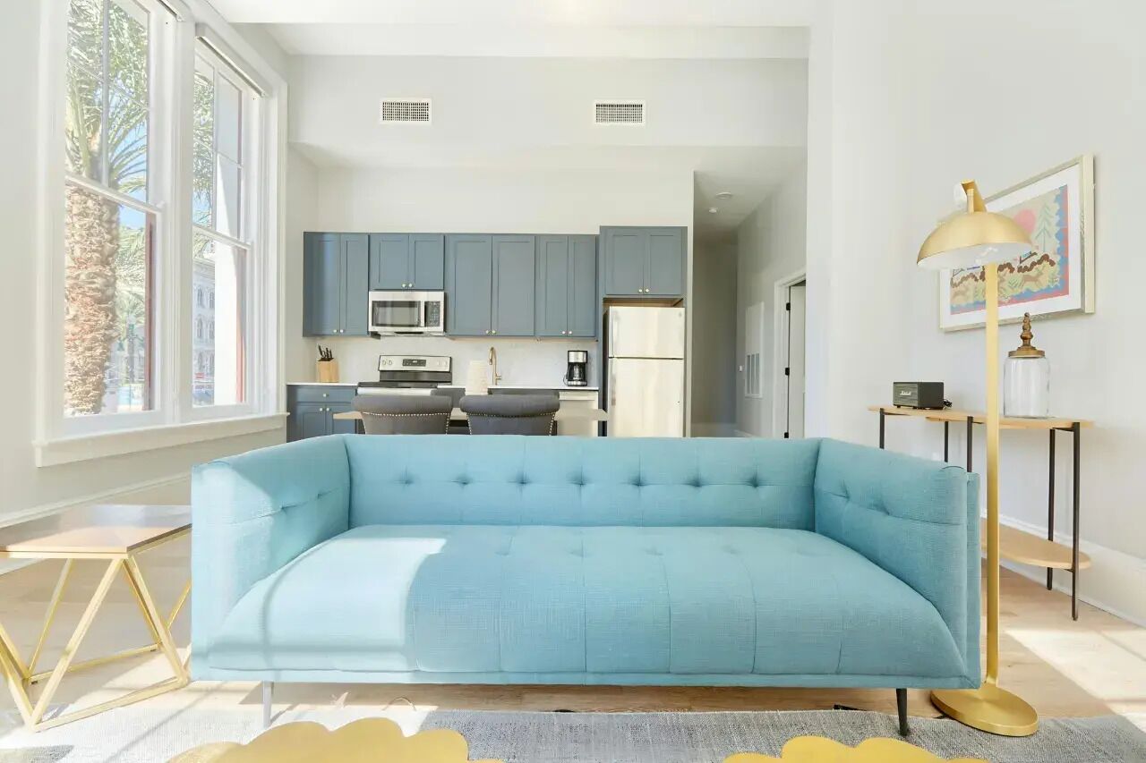 A modern living room with a light blue sofa, a small dining table, and a kitchen in the background.