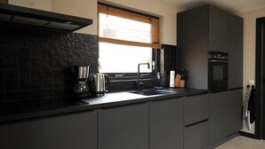 A modern kitchen with dark cabinetry, a black countertop, a coffee maker, a kettle, and an oven. - Fokkemaat - Modern family home with large garden, close to the beach and town center (Callantsoog)