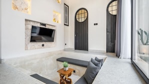 A modern living room with a flat-screen TV mounted on a stone wall, a sofa with cushions, a small wooden table with a plant, and two large black doors. - Luxe Private Pool Villa Santisya 4, 4min-beach by Orivista (Mengwi)