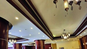 Un hall d’entrée spacieux avec un plafond haut, des lustres modernes et différents espaces de détente, notamment des canapés et un pouf à motifs.