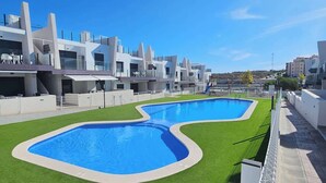 A modern residential complex with a central swimming pool surrounded by artificial grass, white buildings with balconies, and a clear blue sky.