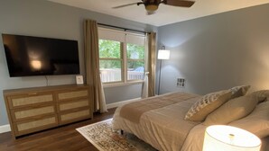 A bedroom with a bed, a TV mounted on the wall, a wooden dresser, a window with curtains, and a ceiling fan.
