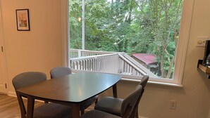 A dining area with a table and four chairs, a window with a view of a balcony and trees, and a framed picture on the wall.