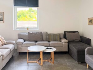 Ein Wohnzimmer mit einer Couch, zwei Beistelltischen, einem Fenster mit Jalousien und einer Zimmerpflanze.