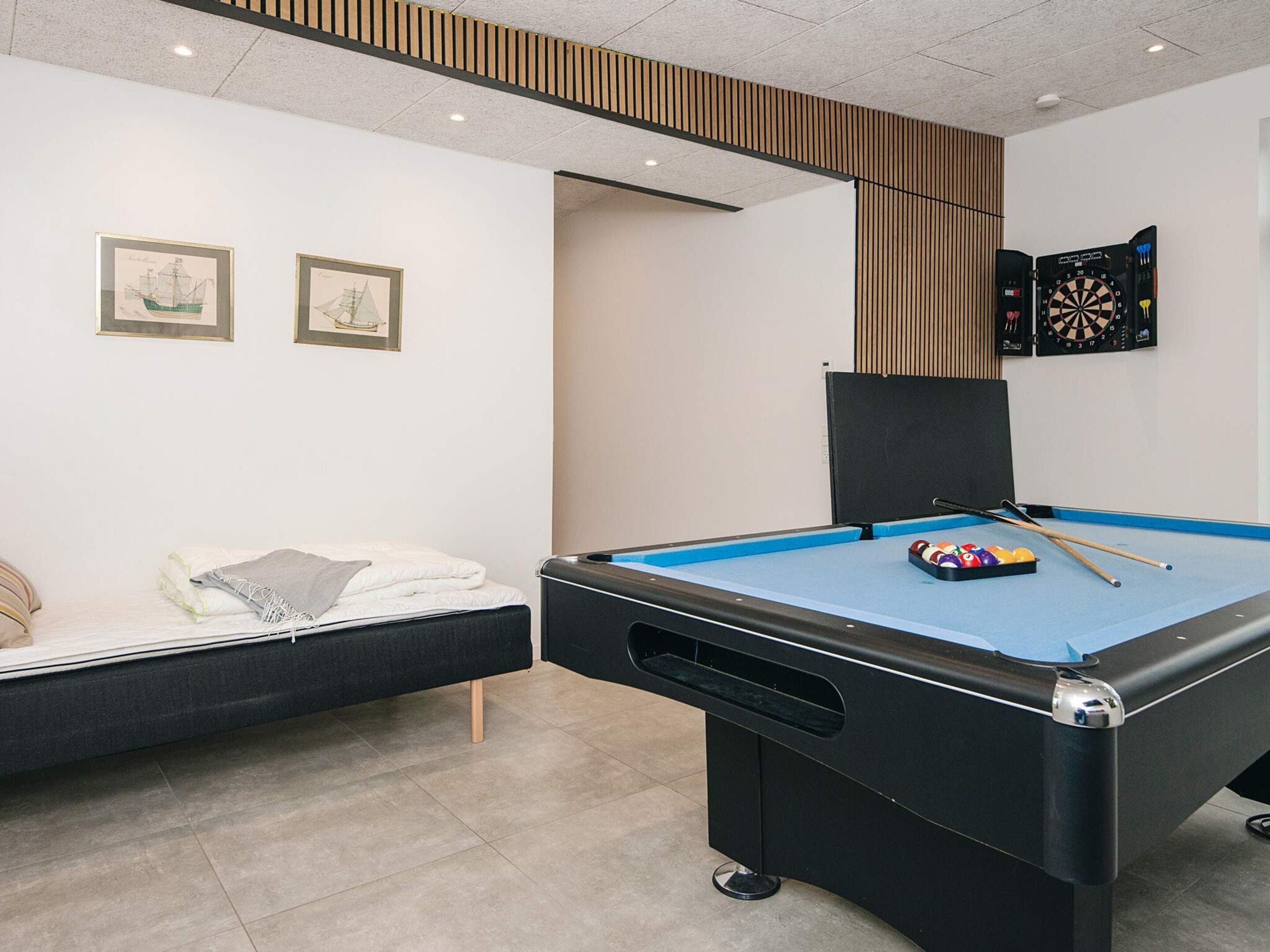 Ein Billardtisch mit blauer Spielunterlage, eine Reihe Billardstöcke und eine Ballpyramide. Ein Bett mit weißer Decke und Kissen, und zwei gerahmte Bilder an der Wand.