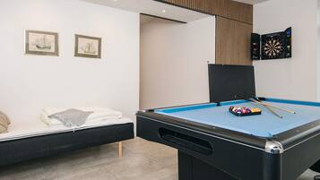 Ein Billardtisch mit blauer Spielunterlage, eine Reihe Billardstöcke und eine Ballpyramide. Ein Bett mit weißer Decke und Kissen, und zwei gerahmte Bilder an der Wand.