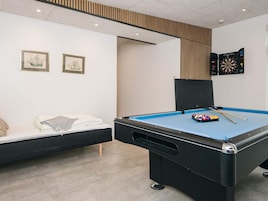 A pool table with a blue felt surface, a set of pool cues, and a rack of balls. There is a bed with a white comforter and pillows, and two framed pictures on the wall.