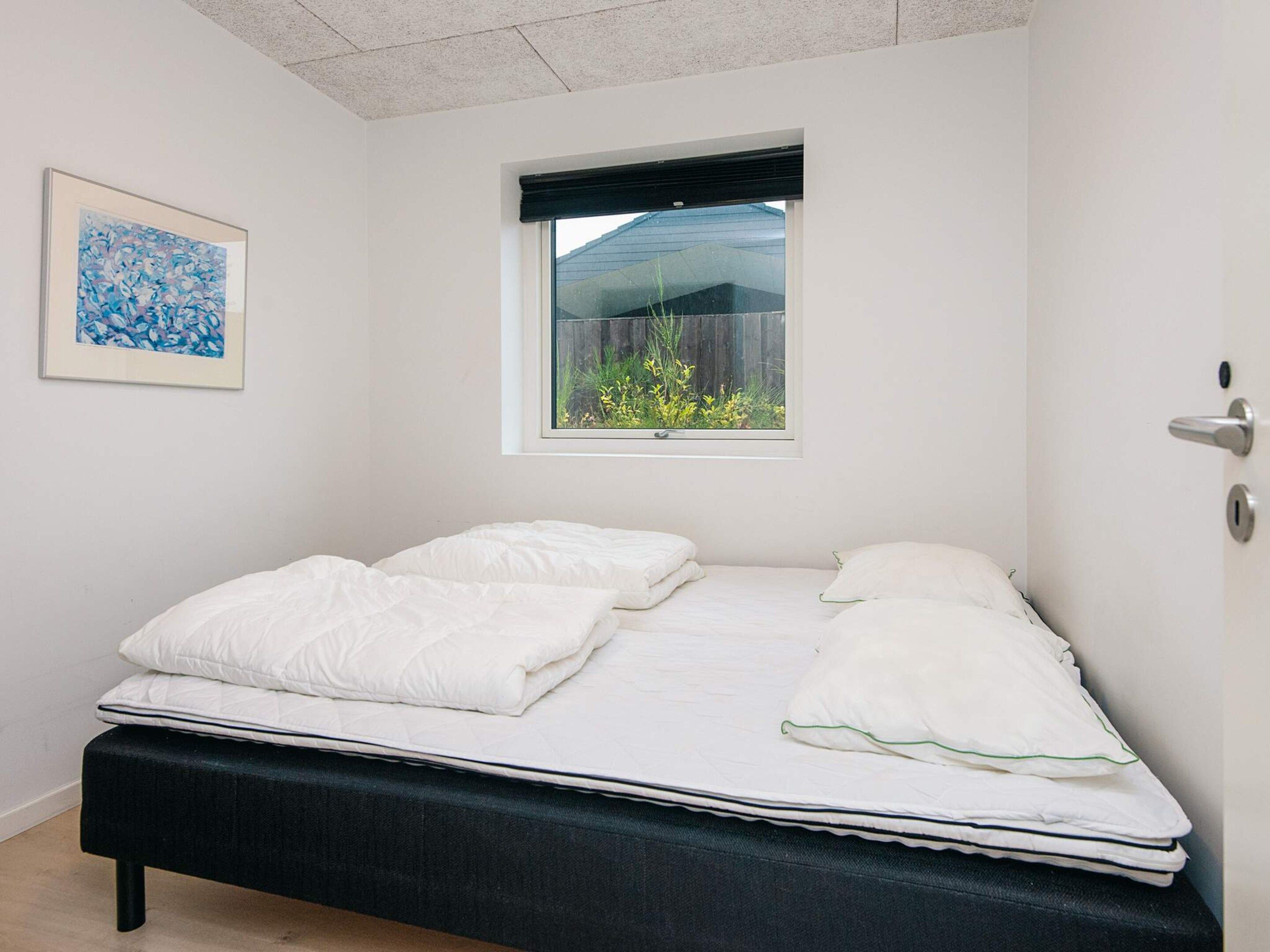 A neatly made bed with white bedding in a room with white walls and a window with a view of greenery.
