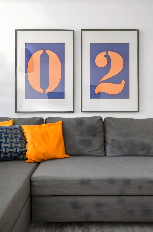 A grey sofa with two framed artworks above it, a blue patterned pillow, and an orange pillow.