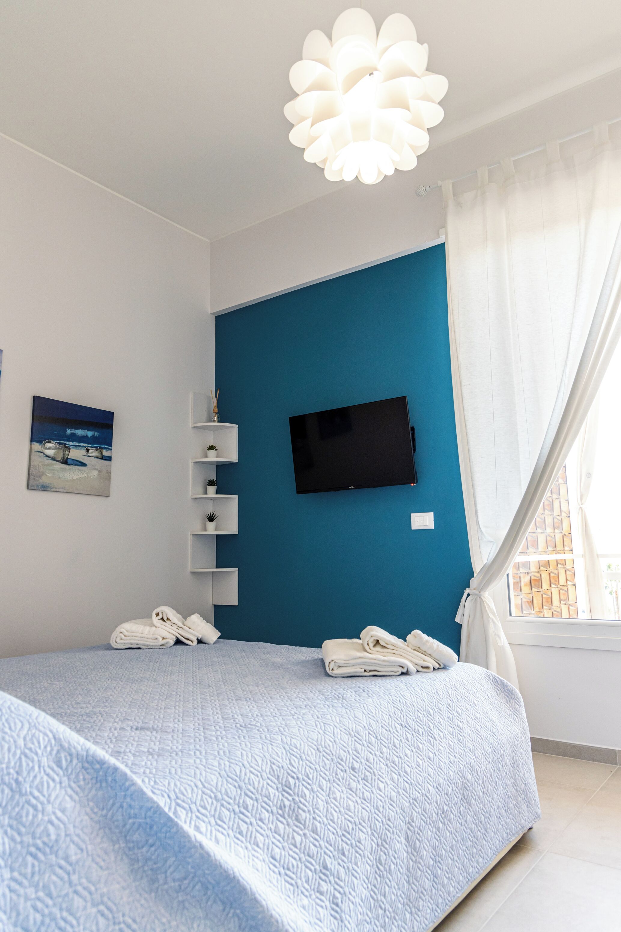 A bedroom with a bed, a TV mounted on the wall, a window with curtains, and a beach-themed painting on the wall.
