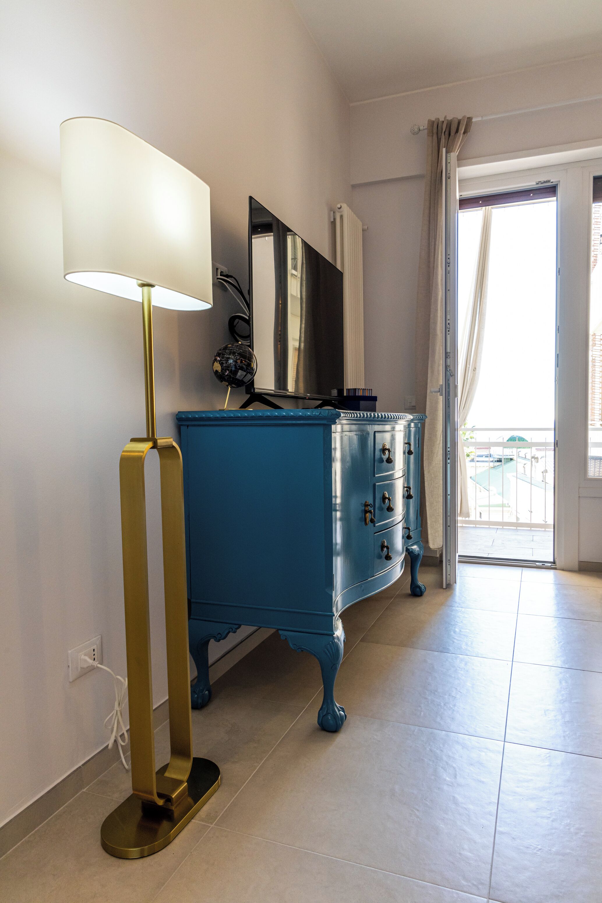 A room with a blue dresser, a standing lamp, a mirror, and a view of a balcony.