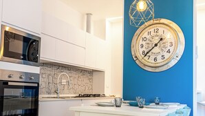 A modern kitchen with white cabinetry, a blue accent wall, a round clock, and a dining area with white and blue chairs. - Casinò Charme with Terrace by Wonderful Italy (Sanremo)