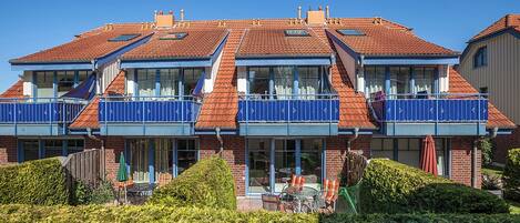 Un bâtiment à deux étages, doté d’un toit recouvert de tuiles rouges, de balcons avec des rampes bleues et d’un patio aménagé avec du mobilier d’extérieur.