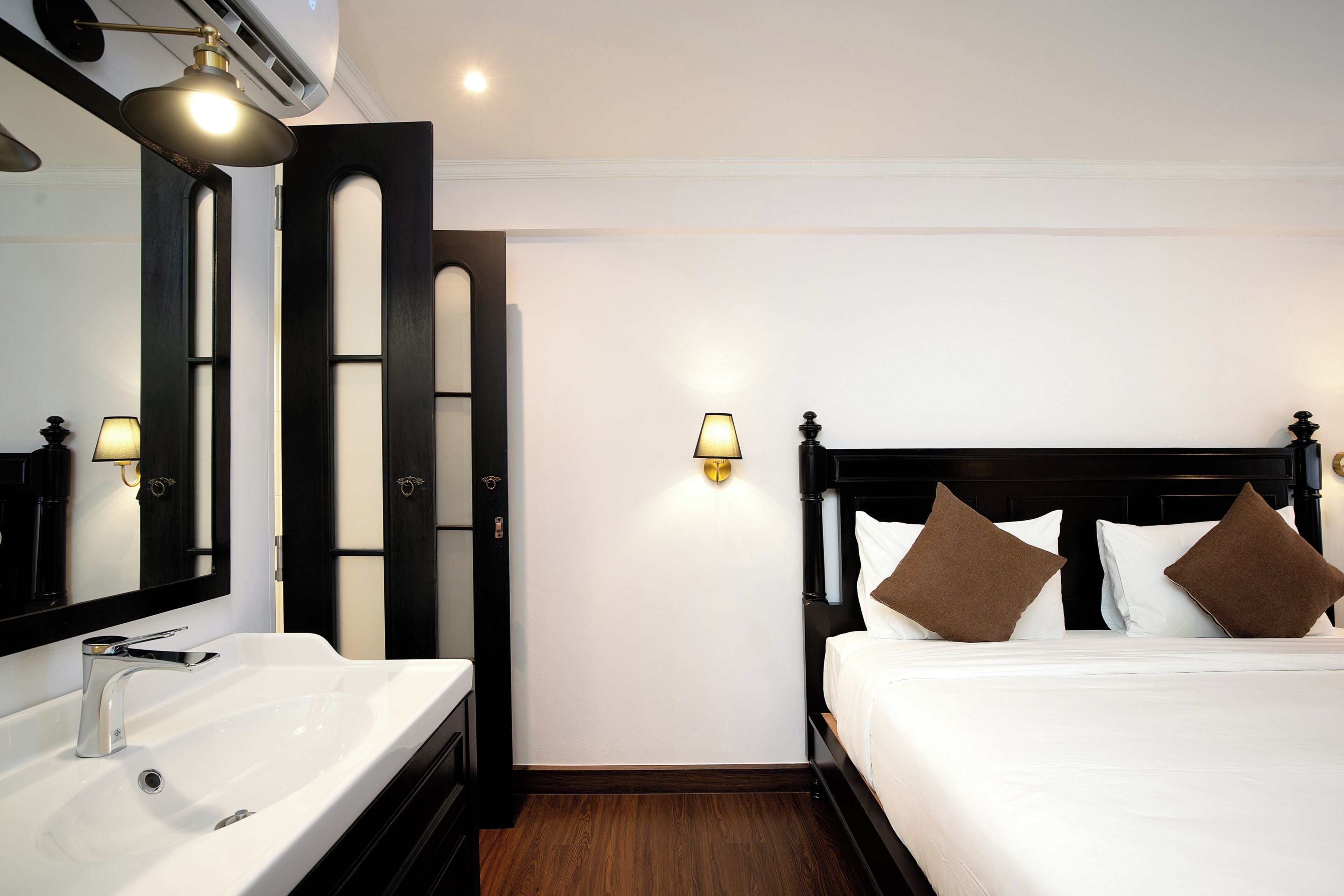 Habitación de hotel con un lavamanos blanco, un espejo con marco negro, una cama con ropa de cama blanca y almohadas marrones, y un aplique de pared.