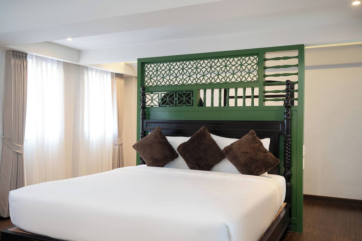 Une chambre d’hôtel avec un lit, des rideaux, un paravent décoratif vert et une tête de lit en bois.