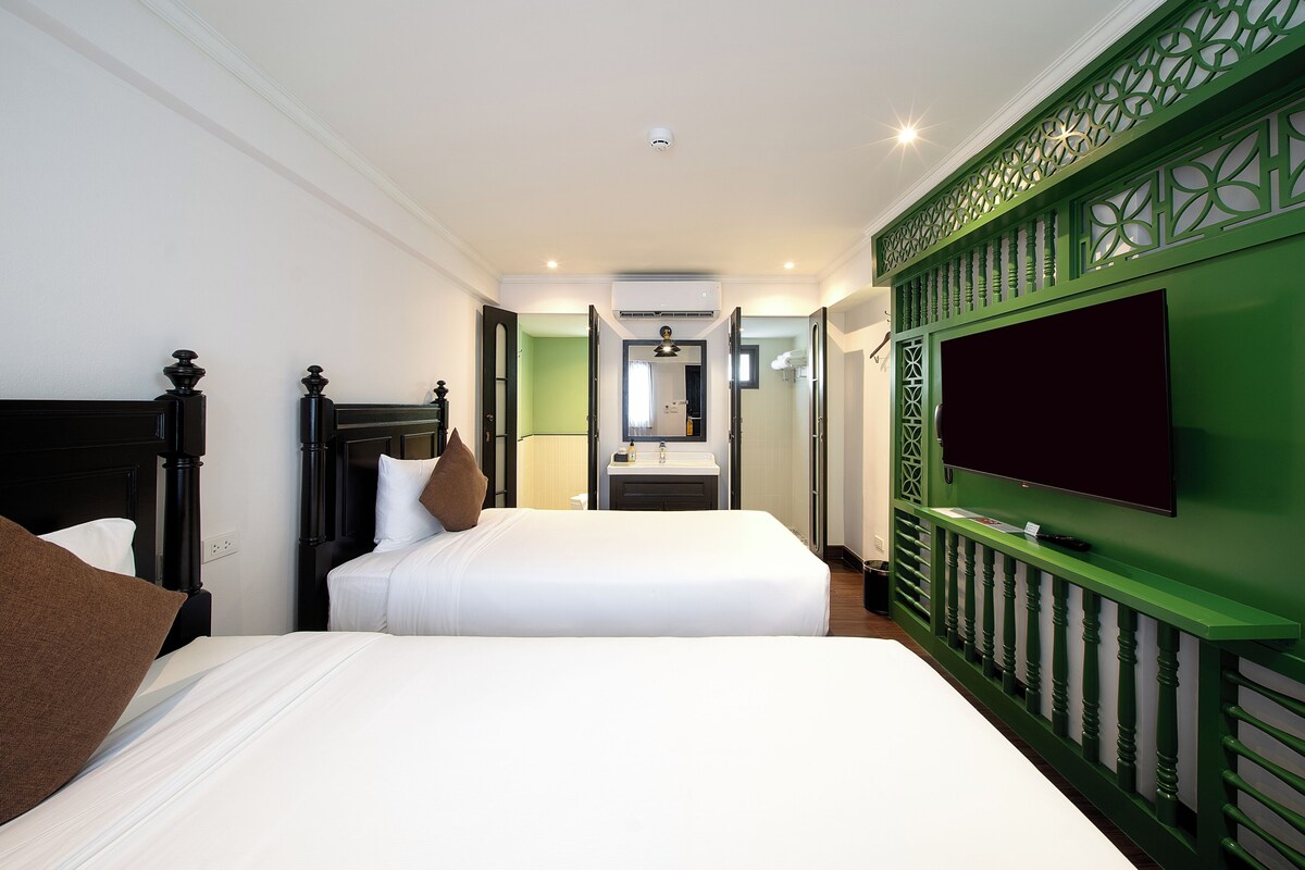 Habitación de hotel con dos camas, un televisor montado en una pared verde, un baño visible a través de una puerta abierta y una cámara de seguridad en el techo.