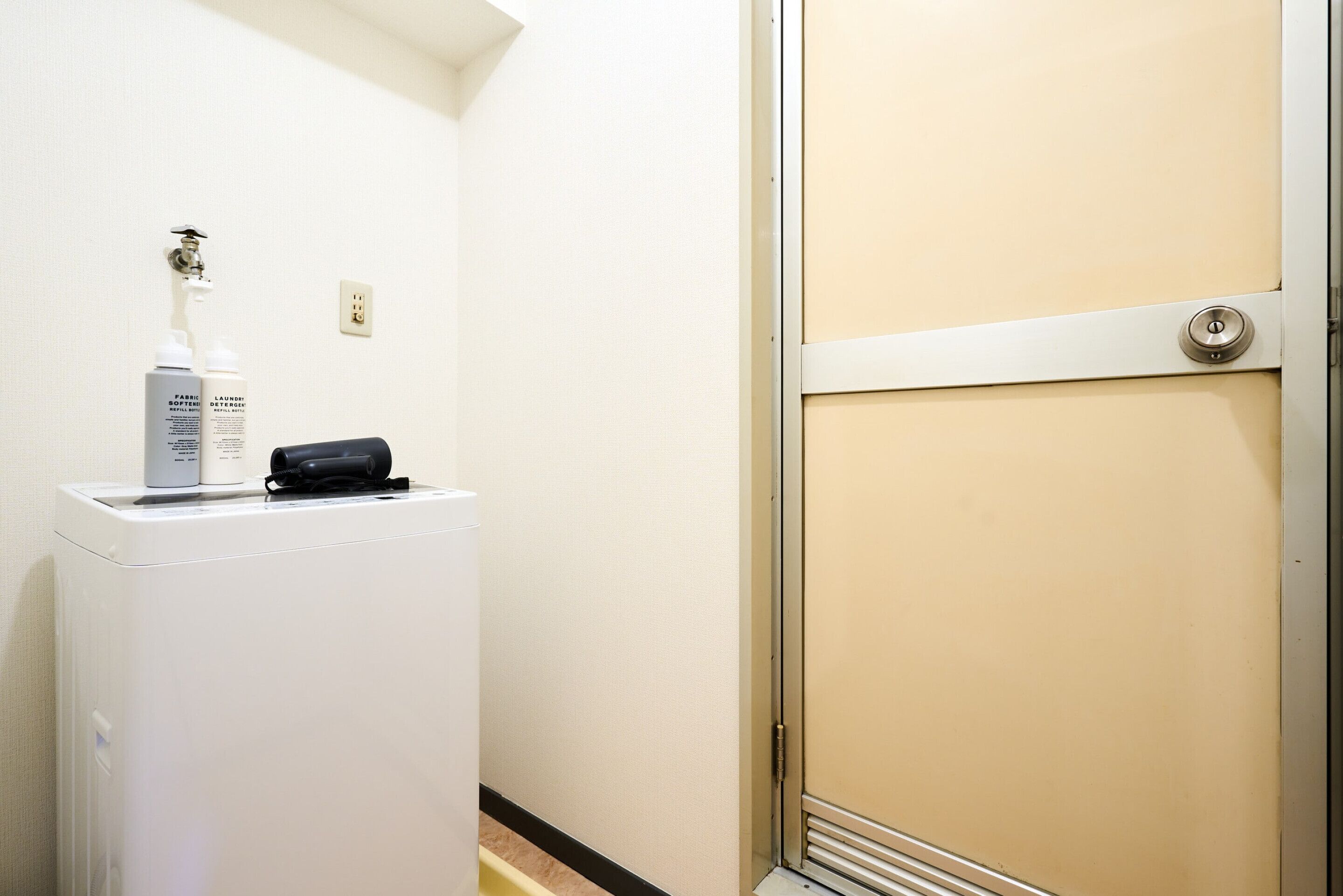 Une chambre d’hôtel avec un mini-bar blanc, un sèche-cheveux noir et une porte beige.
