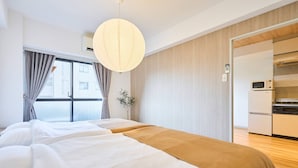 A hotel room with a bed, a hanging light fixture, a window with curtains, and a kitchenette area.