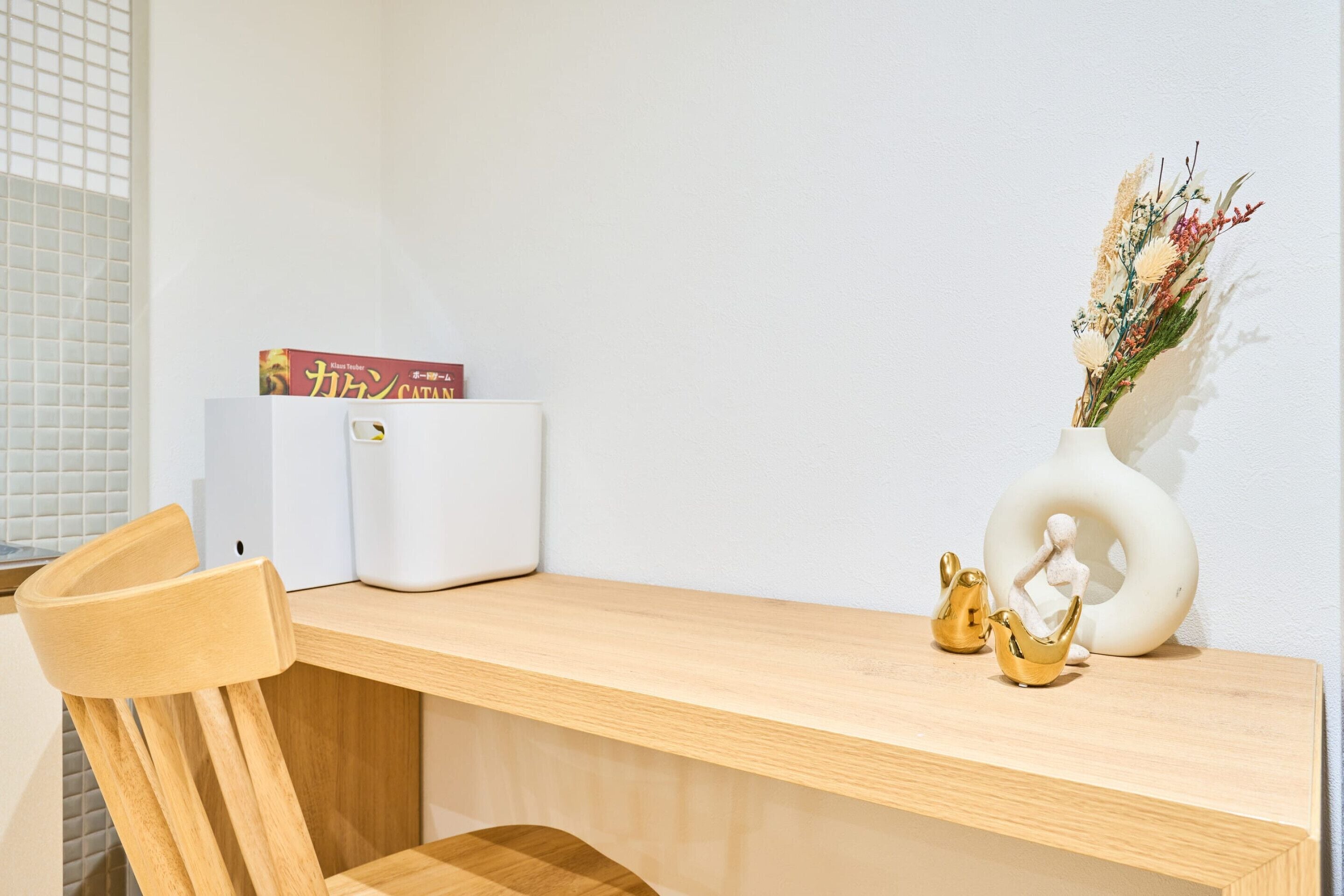 Ein Holztisch mit einer weißen Vase voller getrockneter Blumen, einer kleinen goldenen Figur und einem Buch mit dem Titel 'Catan'.