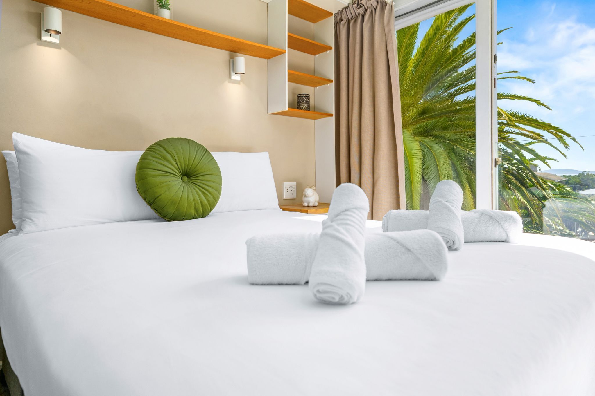 Un lit bien fait, avec du linge de lit blanc et un coussin vert en guise d’élément décoratif, une table de chevet agrémentée d’un petit objet décoratif, et une vue sur des palmiers à travers une fenêtre ornée de rideaux.