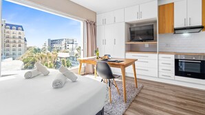 A modern hotel room with a large bed, a dining area with a table and chairs, a kitchenette with a TV and oven, and a view of palm trees and buildings. - Greenpark 202 (Cape Town)