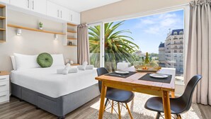A modern bedroom with a large bed, a dining table, and a view of palm trees and buildings. - Greenpark 202 (Cape Town)
