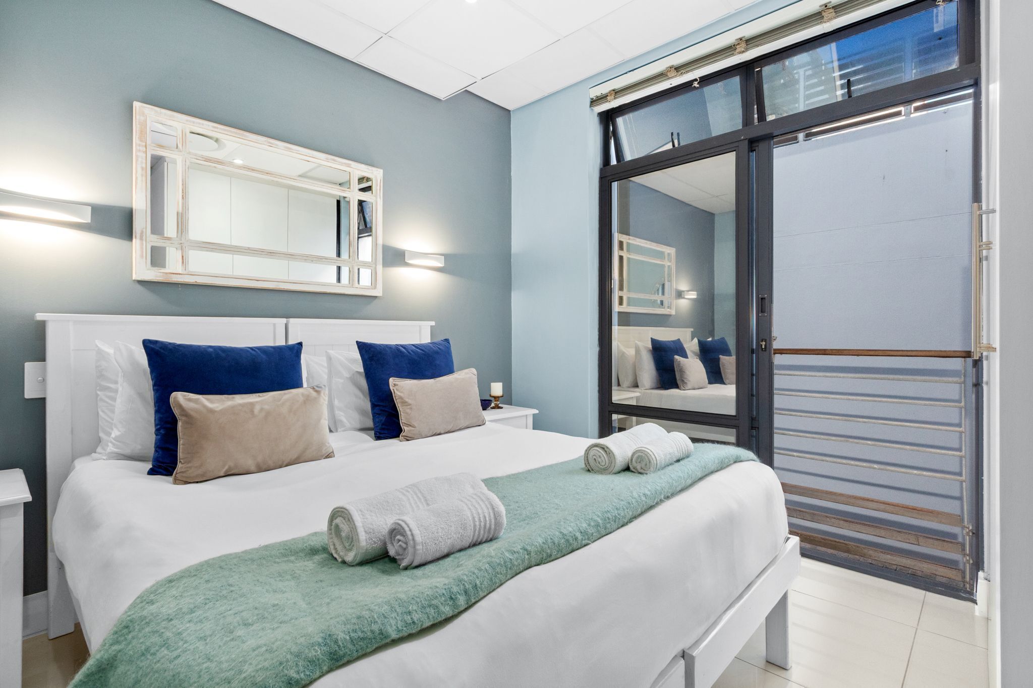 A neatly made bed with a green blanket and rolled towels, a large mirror above it, and a sliding glass door to a balcony.