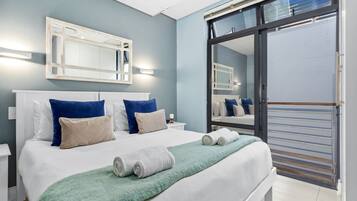 A neatly made bed with a green blanket and rolled towels, a large mirror above it, and a sliding glass door to a balcony.