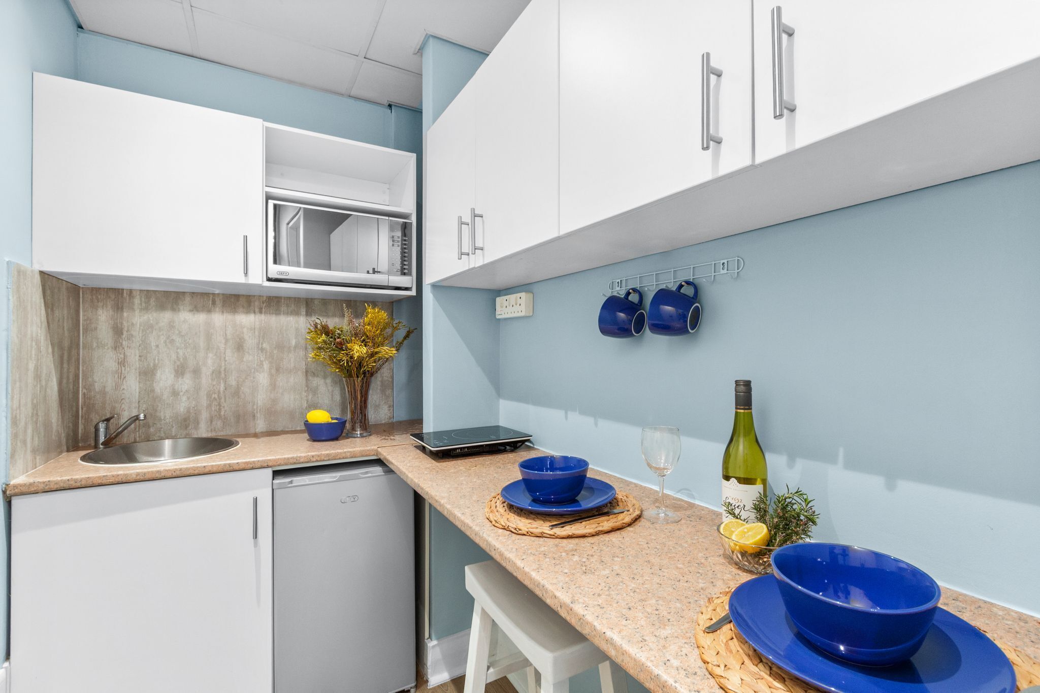A modern kitchen with white cabinets, a stainless steel sink, and a built-in microwave. There are blue bowls on the counter, a wine bottle, and a small plant.